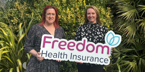 Two women holding a 'Freedom Health Insurance' sign outdoors, smiling amid green foliage.