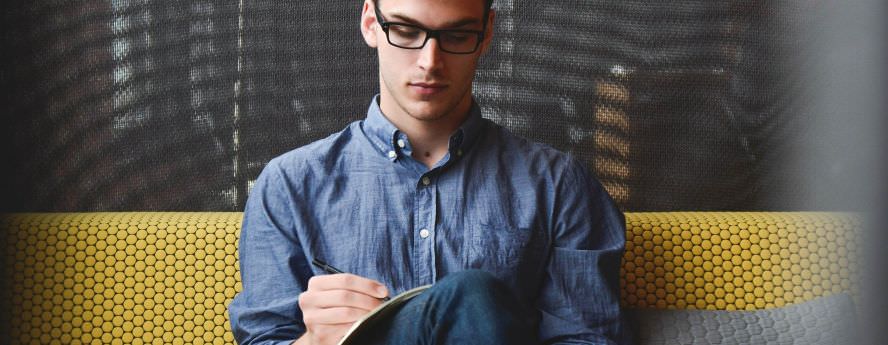 Young Man With Glasses Sitting On Couch And Taking Notes Studying 