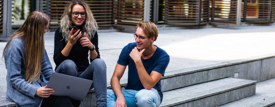 Group Of Young People Sitting On University Building Stairs Talking Studying And Browsing On Laptop